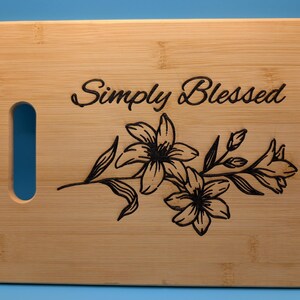 May include: A rectangular bamboo chopping board with a handle cutout on the left. The words "Simply Blessed" are above a floral lily design. The text and floral design are in black.