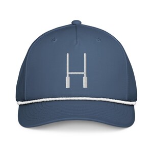 Op de afbeelding: Een marineblauwe baseballcap met een wit touwdetail langs de rand. De cap heeft een wit geborduurd "H"-ontwerp op de voorkant. Een modieus accessoire.