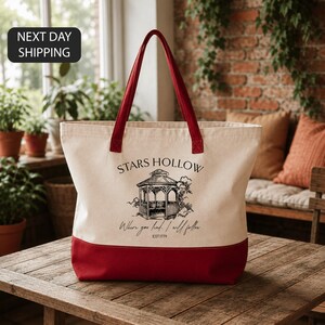 A beige tote bag with a red trim, featuring a graphic of a gazebo and the text "STARS HULLOW" and "Where you find me I will follow" printed on it. The bag is placed on a wooden table, with a brick wall and potted plants in the background.