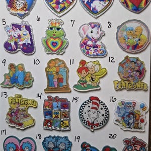May include: A collection of twenty numbered acrylic charms featuring various cartoon characters, including The Flintstones, Dr. Seuss figures, and animals. Each charm has a distinct shape, colorful graphic prints, and a glittery border finish.