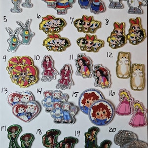 May include: A collection of twenty numbered acrylic charms featuring various cartoon characters, pop culture figures, and animals. Each charm has a glittery or colored border, including designs of SpongeBob characters, Powerpuff Girls, Selena, cats, Raggedy Ann, Princess Peach, Woody from Toy Story, and coffee cups.