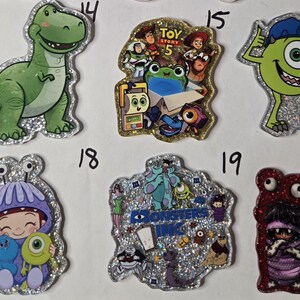 May include: A collection of six glittery acrylic character pins featuring Rex, a Toy Story 5 collage, Mike Wazowski with a clipboard, a child in a purple monster costume, a Monsters Inc. logo collage, and a scene with Boo, Sulley, and Mike.