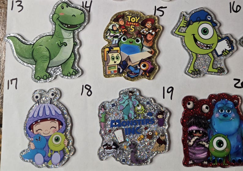 May include: A collection of six glittery acrylic character pins featuring Rex, a Toy Story 5 collage, Mike Wazowski with a clipboard, a child in a purple monster costume, a Monsters Inc. logo collage, and a scene with Boo, Sulley, and Mike.