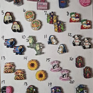 May include: A collection of twenty pairs of small acrylic charms featuring various colorful graphic designs, including cartoon characters, animals, flowers, and text-based slogans, arranged in a numbered grid on a plain white background.