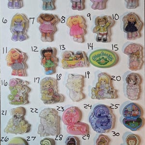 May include: A collection of vintage Cabbage Patch Kids and Precious Moments stickers and charms. The stickers feature various dolls in different outfits. Also included are miniature toy sets.