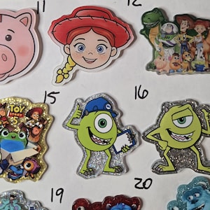 May include: A collection of acrylic character charms featuring various animated figures including a pink pig, a cowgirl, a group of toys, a green one-eyed monster wearing a blue hat, and a green monster pointing, with some charms having glitter.