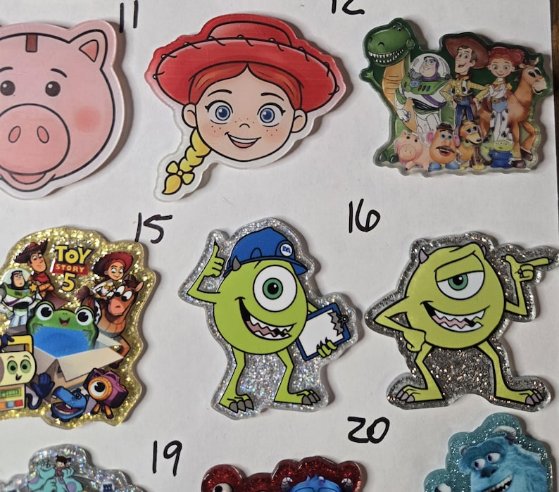 May include: A collection of acrylic character charms featuring various animated figures including a pink pig, a cowgirl, a group of toys, a green one-eyed monster wearing a blue hat, and a green monster pointing, with some charms having glitter.