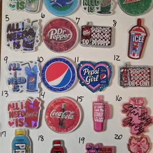 May include: A collection of twenty numbered acrylic beverage-themed charms featuring various soda and drink brands including Sprite, Dr. Pepper, Mountain Dew, Icee, Pepsi, Coca-Cola, Stanley, Prime, and Alani, with some designs incorporating bows and text.