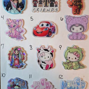 May include: A collection of twelve colorful, holographic charms. The charms feature various designs, including cartoon characters, horror movie characters, and baby shower announcements. The word "FRIENDS" is visible on one charm, and "IT'S A GIRL" and "IT'S A BOY" are on others.