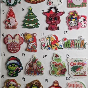 May include: A collection of twenty numbered acrylic Christmas-themed charms featuring various designs including a sweater, bells, a ghost, a snowman, a gnome, a tree, a reindeer, Snoopy, Minnie Mouse, Winnie the Pooh, and Grinch characters.