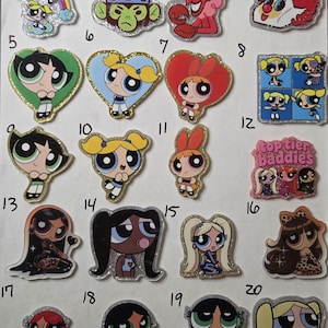 May include: A collection of twenty numbered acrylic pins featuring various cartoon characters, including the Powerpuff Girls, Mojo Jojo, Him, and a red telephone. Each pin has a glittery finish and depicts colorful, stylized character illustrations.