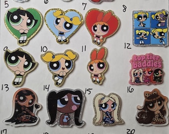 May include: A collection of twenty numbered acrylic pins featuring various cartoon characters, including the Powerpuff Girls, Mojo Jojo, Him, and a red telephone. Each pin has a glittery finish and depicts colorful, stylized character illustrations.