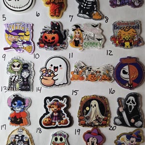 May include: A collection of twenty numbered acrylic charms featuring various Halloween-themed characters, including ghosts, pumpkins, and cartoon figures, with glittery finishes and printed designs on a white background.