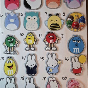 May include: A collection of twenty colorful, glitter-edged charms. The charms feature various designs, including a strawberry, cow, cat, axolotl, unicorn, owl, bee, M&M's characters, and Miffy the bunny in different outfits. The charms are made of plastic.