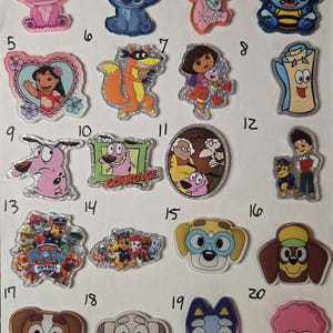 May include: A collection of twenty numbered acrylic character charms featuring various animated figures including Stitch, Lilo, Dora the Explorer, Courage the Cowardly Dog, Paw Patrol characters, and Bluey-themed designs with glitter and clear finishes.