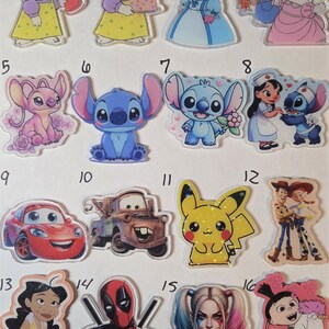 May include: A collection of sixteen numbered acrylic character charms featuring various animated figures including rabbits, aliens, a yellow electric creature, a red race car, a tow truck, a superhero, a clown, and a girl with a unicorn.