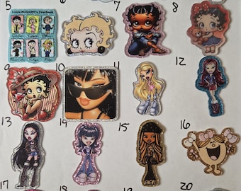 May include: A collection of twenty numbered acrylic character charms featuring various animated and doll-inspired figures, including Betty Boop, Bratz dolls, Lizzie McGuire, and Wednesday Addams, each with unique glittery borders and distinct graphic designs.