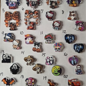 May include: A collection of twenty numbered sets of acrylic charms featuring various Halloween and pop culture characters, including ghosts, skulls, pumpkins, cartoon figures, and movie icons, arranged in a grid on a white background.