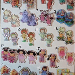 May include: A collection of twenty pairs of acrylic charms featuring various cartoon characters, babies, and dolls, each numbered from 1 to 20. The charms display colorful illustrations of figures in diverse outfits, including some with holographic details and clear borders.