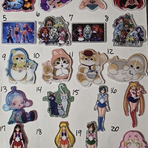 May include: A collection of twenty numbered acrylic charms featuring various anime and cartoon characters, including cats, groups of figures, and individual characters in colorful outfits, all arranged on a white background for display.