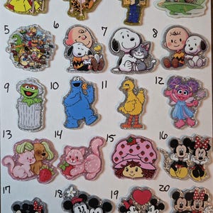 May include: A collection of glittery character pins featuring cartoon characters like Mickey Mouse, Snoopy, and characters from Rugrats and Sesame Street. Each pin has a silver glitter border and is on a white surface.