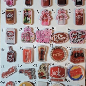 May include: A collection of clear acrylic charms featuring various designs. Includes Dr. Pepper cans and bottles, Coca-Cola logos, and fast-food brand logos like Burger King and Arby's. Some charms have text like "Hot Girls Drink Dr. Pepper".