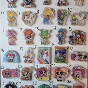 May include: A collection of clear acrylic charms featuring cartoon characters and designs. The charms include images of Rainbow Brite characters, Powerpuff Girls, and Starbucks-themed designs. The charms are in various colors and shapes.