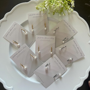 May include: Several pairs of hoop earrings with small white pearls, each mounted on a branded Still We Bloom Shop display card. The earrings feature gold-tone and silver-tone metal hoops. The cards are arranged on a white scalloped plate.
