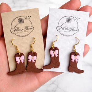 以下が含まれることがあります： ピンクのリボンが付いた茶色のカウボーイブーツのイヤリング。ベージュと白のカードに表示されています。イヤリングには金色のフックが付いています。カードには「Still We Bloom Shop Co.」の文字とタンポポのロゴがあります。