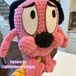 May include: A pink crocheted puppy plushie with the text "Cowardly Pink Puppy" at the top. The puppy has large white eyes with black pupils, black ears, and a black nose. The text "Pattern By: @glitterbeecreations" is also visible.