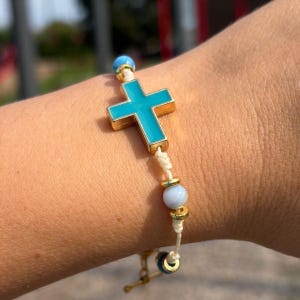 May include: A bracelet featuring a central turquoise-coloured cross charm with a gold-tone border, accented by light blue spherical beads and small gold-tone spacers, all strung on a thin, off-white cord with a knotted design.