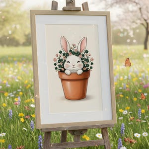 May include: A framed print on an easel features a charming illustration of a white bunny with pink-lined ears, nestled in a terracotta pot surrounded by flowers. The artwork is set against a soft, neutral background, evoking a spring theme.