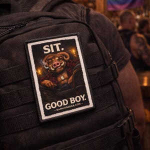 May include: Black backpack with a rectangular patch showing a cartoon boar in a plaid shirt, smoking. The patch reads "SIT. GOOD BOY." and "-RAMHOGGEAR.COM-". A rainbow flag is visible in the background.