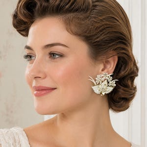 May include: A close-up of a vintage-style earring. The earring features a floral design with white and gold accents, and is adorned with small, sparkling crystals. The earring is clipped to the ear.