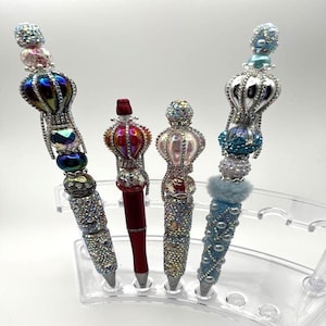 May include: Four decorative pens with ornate, jewel-encrusted designs. The pens feature a variety of colors, including silver, red, blue, and iridescent accents. Each pen has a unique crown-like topper and is displayed in a clear pen holder.