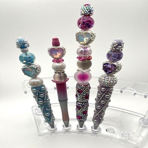 May include: Four decorative pens with beaded and jeweled designs. The pens feature various colors, including blue, pink, and purple, with heart-shaped and round embellishments. The pens are displayed in a clear plastic holder.