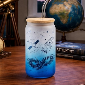 May include: A frosted glass tumbler with a bamboo lid and a clear glass straw. The tumbler features a blue and white gradient design with illustrations of a satellite, a rocket, and swirling galaxies. The theme is astronomy.