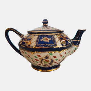 Könnte beinhalten: Dekorative Keramik-Teekanne mit Deckel. Die Teekanne hat eine weiße Basis mit blauen, roten und goldenen Akzenten. Henkel und Ausguss sind dunkelblau mit goldenen Verzierungen. Traditionelles Design.