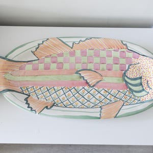 Puede incluir: Una fuente decorativa con forma de pez y un dise&ntilde;o colorido. El pez presenta un patr&oacute;n a cuadros en rosa y verde, rayas y una cabeza punteada. La fuente est&aacute; sobre una superficie blanca y tiene un borde verde.