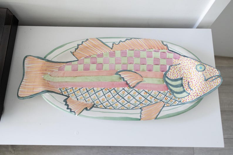 Puede incluir: Una fuente decorativa con forma de pez y un dise&ntilde;o colorido. El pez presenta un patr&oacute;n a cuadros en rosa y verde, rayas y una cabeza punteada. La fuente est&aacute; sobre una superficie blanca y tiene un borde verde.