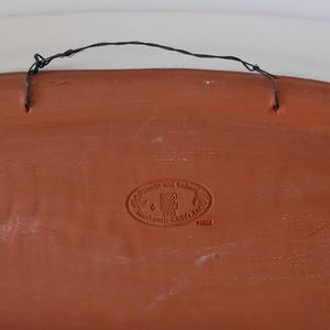 Puede incluir: Parte trasera de un plato decorativo de color terracota con un alambre negro para colgar. El plato tiene un emblema estampado que dice "Victoria and Richard Mackenzie-Childs, Ltd. 1983".