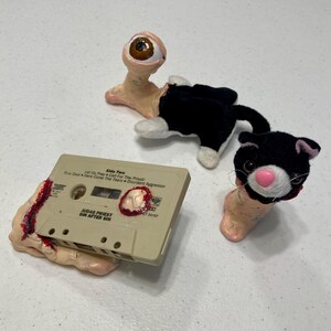 Könnte beinhalten: Eine künstlerische Anordnung mit einer Kassette, einem schwarz-weißen Katzenplüsch und einer Kreatur mit einem Augapfel. Auf der Kassette steht "JUDAS PRIEST SIN AFTER SIN". Die Objekte befinden sich auf einer weißen Oberfläche.