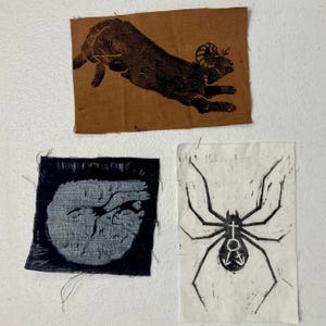 May include: Three fabric patches with printed designs. One patch is brown with a black ram leaping. Another is dark blue with a light gray circular design. The third is white with a black spider graphic.