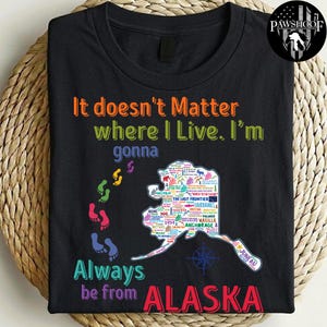 May include: A black t-shirt featuring the text "It doesn't Matter where I Live. I'm gonna Always be from ALASKA" in colourful letters. A map of Alaska filled with city names and icons sits next to a trail of colourful footprints on the left side.