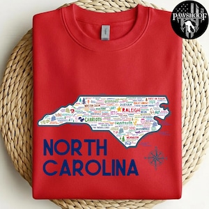 Puede incluir: Una sudadera roja con un mapa de Carolina del Norte lleno de nombres de ciudades. Las palabras "NORTH CAROLINA" est&aacute;n impresas en letras azules grandes debajo del mapa. Un dise&ntilde;o de br&uacute;jula est&aacute; en la parte inferior derecha.