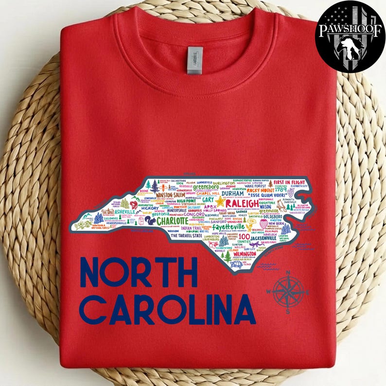 Puede incluir: Una sudadera roja con un mapa de Carolina del Norte lleno de nombres de ciudades. Las palabras "NORTH CAROLINA" est&aacute;n impresas en letras azules grandes debajo del mapa. Un dise&ntilde;o de br&uacute;jula est&aacute; en la parte inferior derecha.