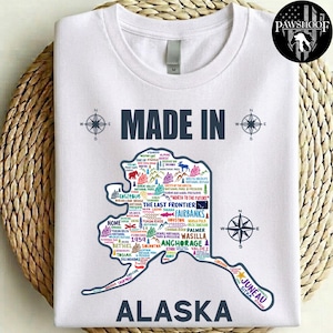 May include: A white short-sleeved t-shirt featuring a graphic of the state of Alaska filled with colourful text and icons representing various cities and landmarks. The text reads MADE IN ALASKA and includes a compass rose on each side.