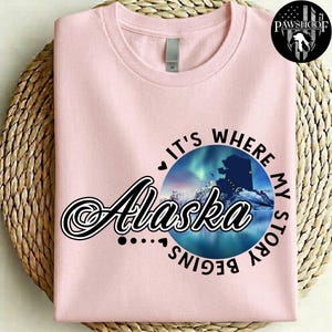 May include: A light pink t-shirt featuring a circular graphic of the Alaska state silhouette against a backdrop of the aurora borealis. The text reads "Alaska" in a large script font, surrounded by the words "It's where my story begins."
