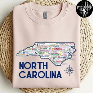 Puede incluir: Una sudadera rosa claro con un contorno detallado de Carolina del Norte, llena de nombres de ciudades y puntos de referencia. Las palabras "NORTH CAROLINA" est&aacute;n impresas en azul marino debajo del mapa, con una rosa de los vientos. Un logotipo est&aacute; en la esquina superior derecha.