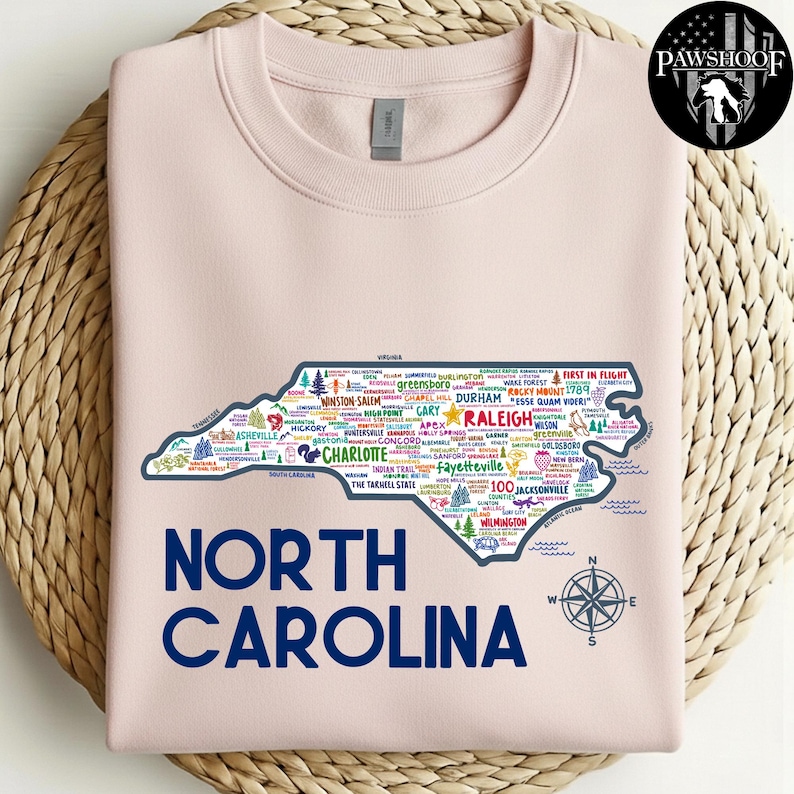 Puede incluir: Una sudadera rosa claro con un contorno detallado de Carolina del Norte, llena de nombres de ciudades y puntos de referencia. Las palabras "NORTH CAROLINA" est&aacute;n impresas en azul marino debajo del mapa, con una rosa de los vientos. Un logotipo est&aacute; en la esquina superior derecha.